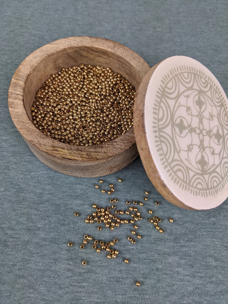 May include: A wooden box with a white lid featuring a decorative design. The box is filled with small, gold-colored beads. Some beads are scattered on a blue surface.