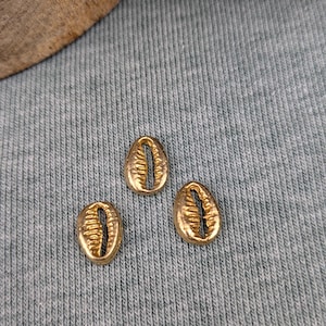 Puede incluir: Tres abalorios de concha de cauri de color dorado sobre una superficie gris texturizada.