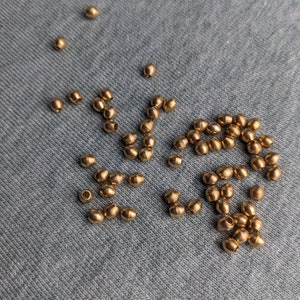 Op de afbeelding: Een verzameling kleine, metallic goudkleurige kralen in een tonvorm met een gladde, reflecterende afwerking. De kralen zijn verspreid over het oppervlak en vertonen een consistente cilindrische vorm met een centraal gat om te rijgen.