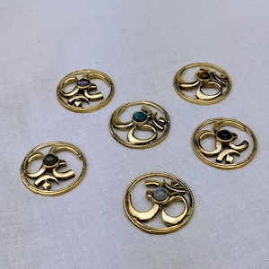 Puede incluir: Seis amuletos Om de color dorado con gemas de diferentes colores en el centro. Los amuletos son redondos y tienen un borde decorativo.