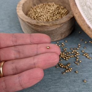May include: A small, gold-colored bead is held in a hand. The bead is very small, and there are many more beads scattered on a blue surface.