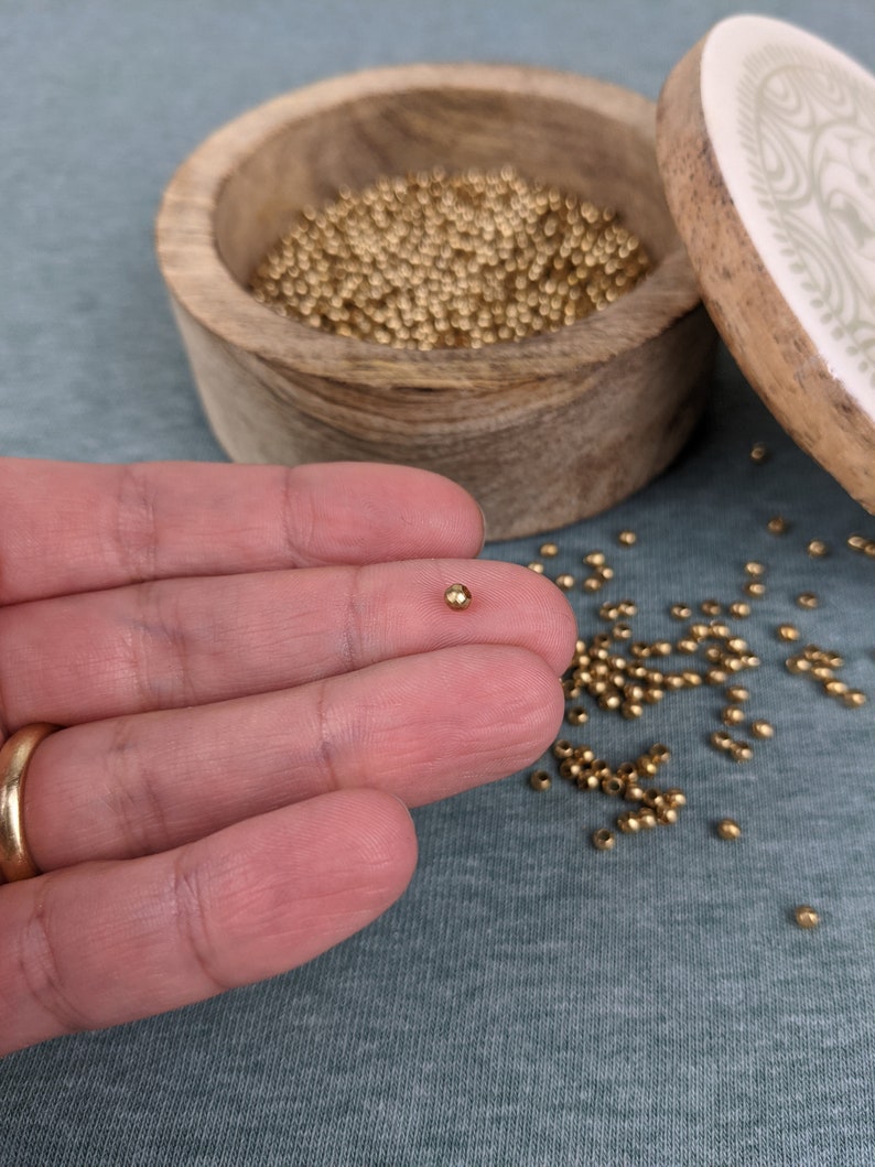 May include: A small, gold-colored bead is held in a hand. The bead is very small, and there are many more beads scattered on a blue surface.