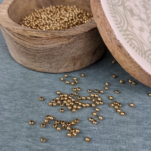 May include: A wooden box with a lid, filled with small gold beads. The lid has a white background with a decorative pattern in brown.