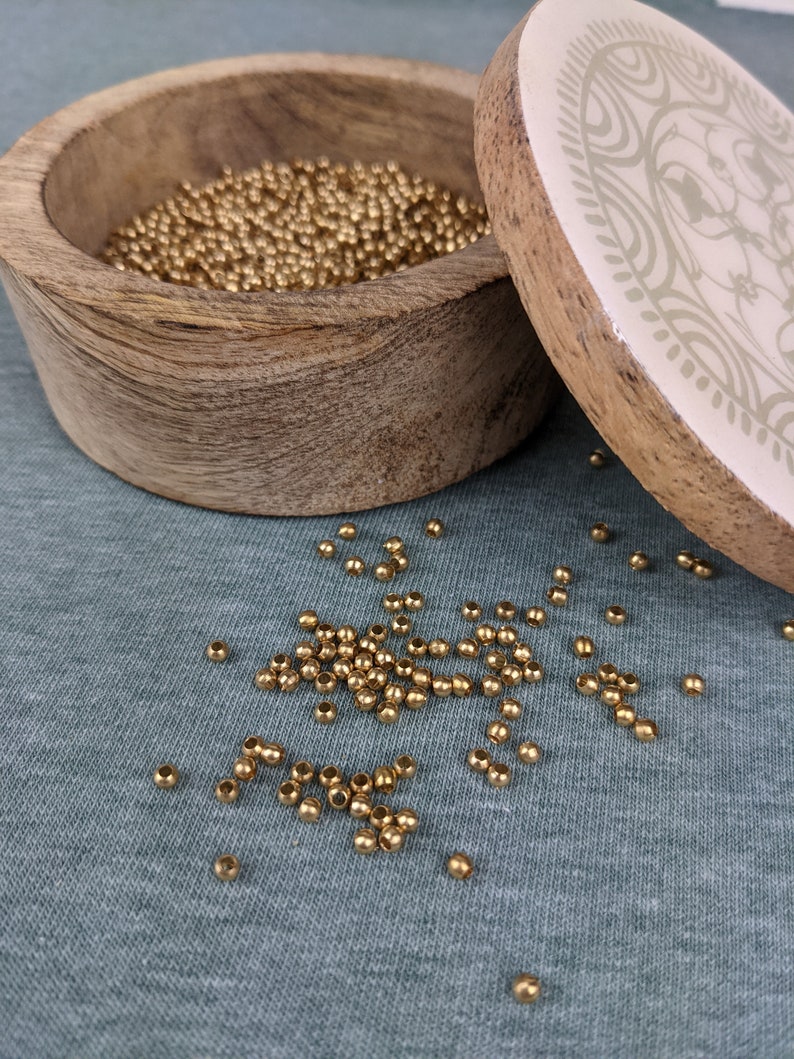 May include: A wooden box with a lid, filled with small gold beads. The lid has a white background with a decorative pattern in brown.