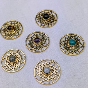 Op de afbeelding: Zes ronde goudkleurige metalen hangers met een ingewikkeld geometrisch sterpatroon. Elke hanger is in het midden voorzien van een andere cabochon edelsteen, waaronder tijgeroog, zwarte onyx, amethist, turkoois, labradoriet en maansteen.