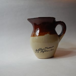 May include: A small, ceramic pitcher with a handle and spout. The top portion is a dark brown, transitioning to a tan middle section, and a cream-colored base. The text "Aztalan Museum Aztalan, Wis" is written on the side.