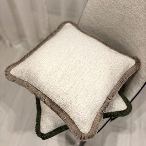 Könnte beinhalten: Zwei quadratische Zierkissen mit cremefarbenem, strukturiertem Stoff und einem Fransenrand. Ein Kissen hat einen braunen Rand, das andere einen grünen Rand. Die Kissen liegen auf einem hellen Stuhl, was auf ein Wohn-Dekor-Thema hindeutet.