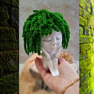 Może przedstawiać: Biała ceramiczna figurka siedzącej osoby z zamkniętymi oczami i zielonymi, teksturowanymi włosami. Figurka trzymana jest w dłoni, widoczna jest złota bransoletka. Tło obejmuje omszałą ceglaną ścianę i drewnianą powierzchnię.