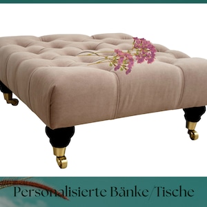 Pode incluir: Um otomano bege claro capitoné com pernas pretas e rodas douradas. O otomano tem uma parte superior quadrada e é decorado com um ramo de flores cor-de-rosa.