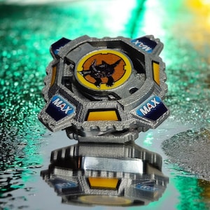 May include: A silver and blue Beyblade with a yellow and orange design. The Beyblade has the word "MAX" printed on it three times. The Beyblade is sitting on a reflective surface.