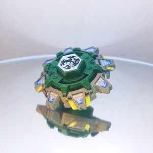 May include: A green and silver Beyblade with a white dragon design on the face. The Beyblade has yellow accents and the words "MAX" and "FIRE" printed on the sides.