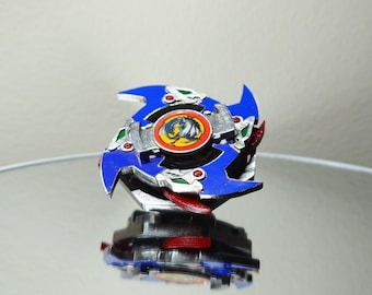 May include: A blue and red Beyblade with a silver and gold centre. The centre has a dragon design and the words "Dragoon" and "Attack" are visible.