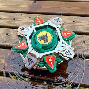 May include: A green and silver toy with a circular design. The toy has a central green piece with a graphic and is surrounded by silver and green components. Red and white accents are visible.