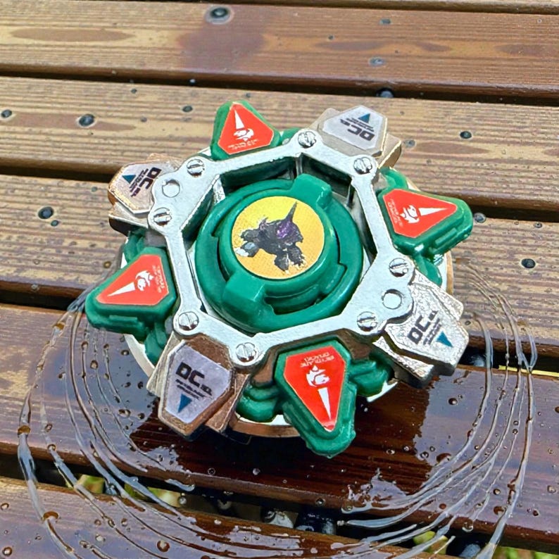 May include: A green and silver toy with a circular design. The toy has a central green piece with a graphic and is surrounded by silver and green components. Red and white accents are visible.