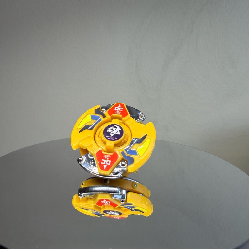 Pu&ograve; includere: Un giocattolo giallo e argento con un design circolare. Il giocattolo ha un emblema centrale e dettagli rossi e blu. Il giocattolo si riflette su una superficie lucida.