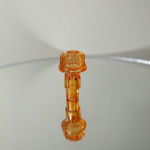 May include: An orange-tinted, decorative bottle stopper. The stopper has a cylindrical body with a detailed, ridged top and bottom. The design is reflected on a mirrored surface, creating a symmetrical image. The stopper is made of a transparent material.