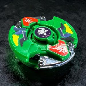 Pu&ograve; includere: Un giocattolo verde e argento con accenti colorati. Il giocattolo ha un design circolare con dettagli rossi, gialli e blu. Il centro presenta un emblema viola con il testo "DRACIEL MF".