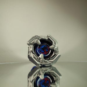 Könnte beinhalten: Ein silberfarbener Kreisel mit einem blau-roten Design in der Mitte. Das Spielzeug hat ein facettenreiches, eckiges Design und spiegelt sich in der glänzenden Oberfläche darunter wider.
