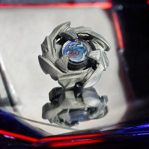 Pode incluir: Um pião prateado e azul com um design de dragão. O pião é feito de metal e tem um centro de plástico transparente.