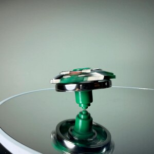 May include: A spinning top toy with a green and silver design. The top is in motion, with a blurred effect indicating its rotation. The base and central stem are green, while the outer ring is silver. The top is set against a reflective surface.
