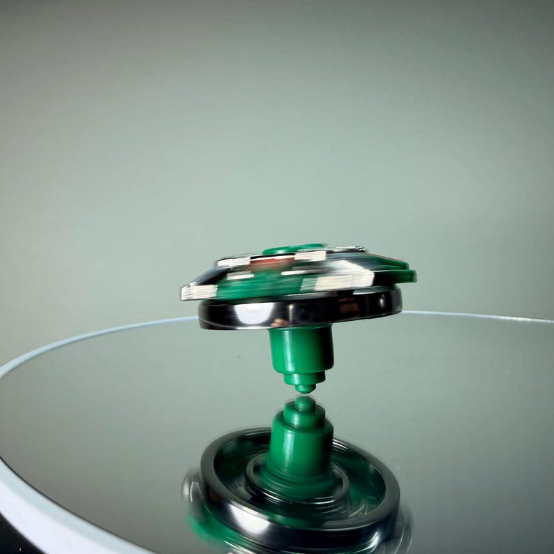 May include: A spinning top toy with a green and silver design. The top is in motion, with a blurred effect indicating its rotation. The base and central stem are green, while the outer ring is silver. The top is set against a reflective surface.