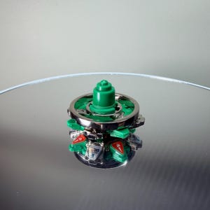 May include: A green and silver spinning top toy. The top has a green central peg, a silver ring, and green and silver blades with red accents. The toy is on a reflective surface.