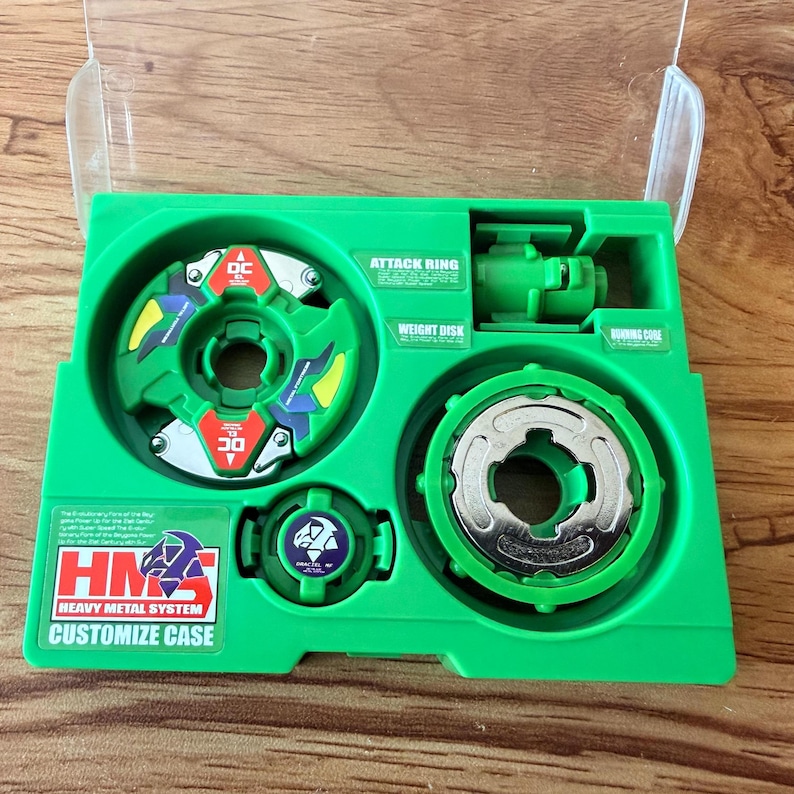 Pu&ograve; includere: Una custodia di giocattoli in plastica verde contenente vari componenti di una trottola. La custodia include un anello d'attacco, un disco di peso e un nucleo di combustione. Il testo "HMS Heavy Metal System Customize Case" &egrave; visibile.