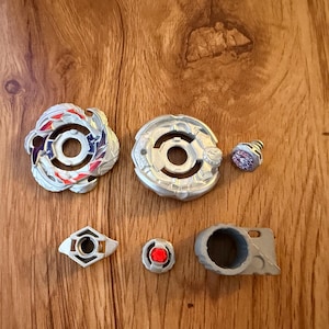 Puede incluir: Colecci&oacute;n de peonzas de juguete y componentes. La imagen muestra dos peonzas principales con dise&ntilde;os intrincados en plata, rojo y azul. Las piezas adicionales incluyen una pieza peque&ntilde;a en forma de cono y otros componentes geom&eacute;tricos.