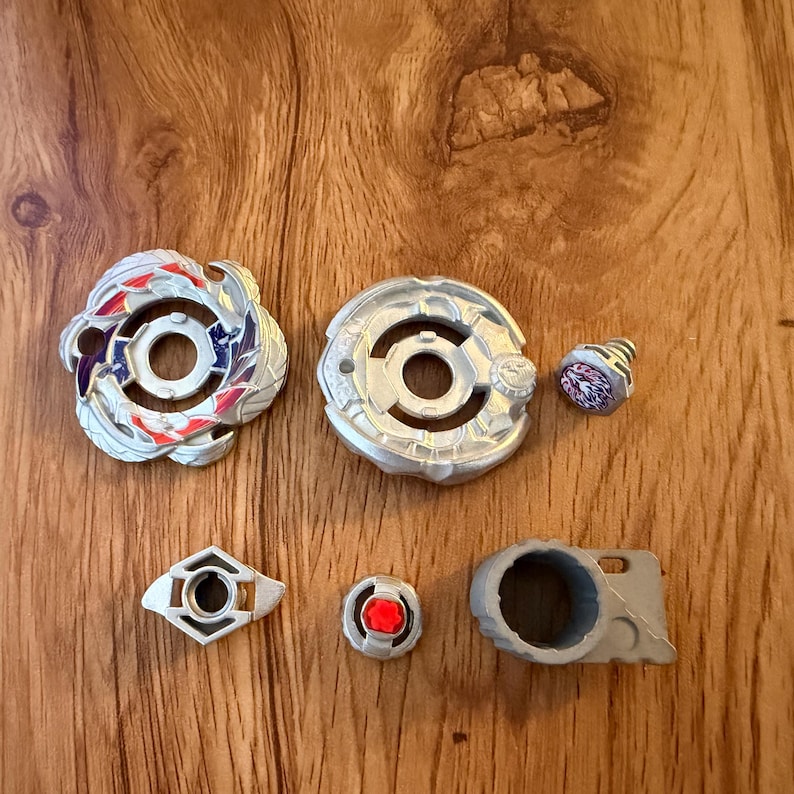 Puede incluir: Colecci&oacute;n de peonzas de juguete y componentes. La imagen muestra dos peonzas principales con dise&ntilde;os intrincados en plata, rojo y azul. Las piezas adicionales incluyen una pieza peque&ntilde;a en forma de cono y otros componentes geom&eacute;tricos.