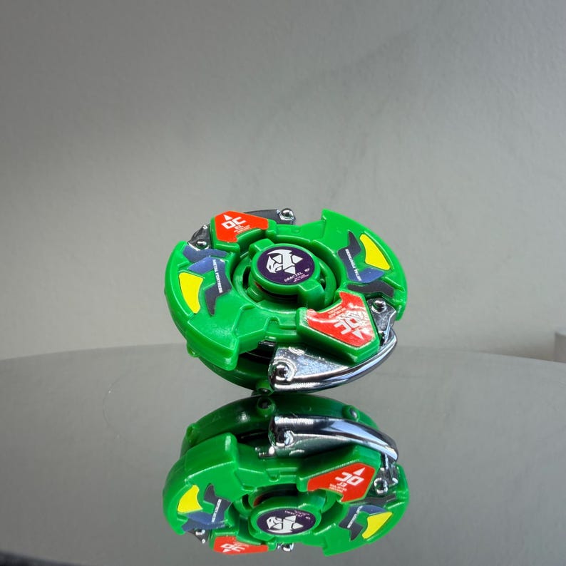 Pu&ograve; includere: Un giocattolo a trottola verde e argento con accenti rossi, gialli e blu. Il giocattolo ha un design circolare con un anello di metallo argentato e un emblema centrale. Il giocattolo si riflette su una superficie lucida.