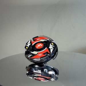 May include: A black and red toy with a circular design, featuring a central emblem and angular accents. The toy has a reflective surface and appears to be a collectible item or a game piece.