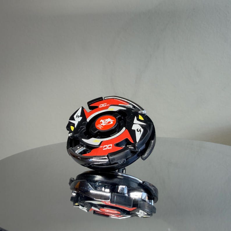 May include: A black and red toy with a circular design, featuring a central emblem and angular accents. The toy has a reflective surface and appears to be a collectible item or a game piece.