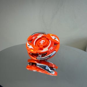 May include: An orange and silver toy with a circular design. The toy has a central orange piece with a silver ring and orange blades. The toy is reflected on a shiny surface. The toy has the number 5 on it.