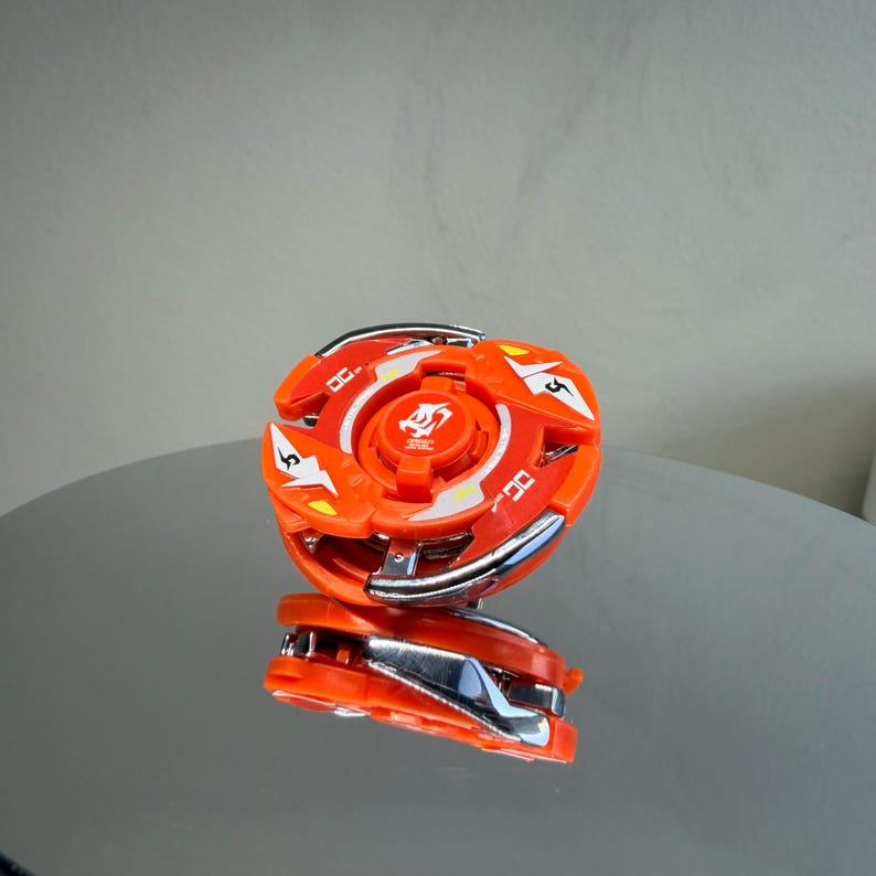 May include: An orange and silver toy with a circular design. The toy has a central orange piece with a silver ring and orange blades. The toy is reflected on a shiny surface. The toy has the number 5 on it.