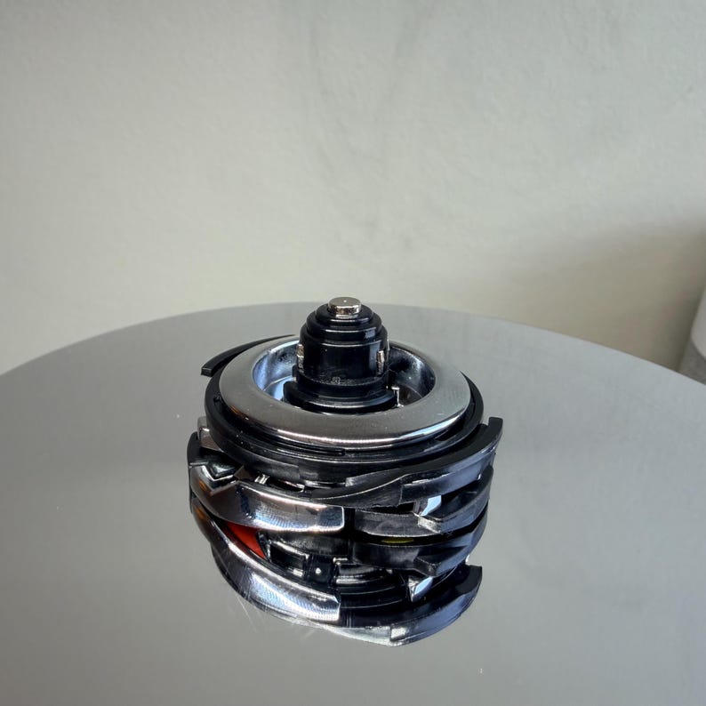 May include: A multi-layered, metallic object with a black top and silver rings. The object has a complex, geometric design with curved and angular elements. Hints of red are visible within the structure, adding a pop of color to the otherwise monochromatic piece.