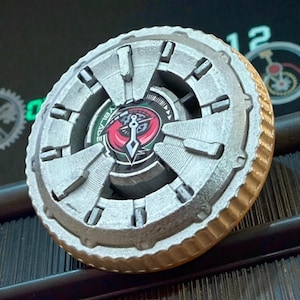 Puede incluir: Un juguete Beyblade plateado y dorado con un emblema rojo y negro en el centro. El juguete tiene un diseño circular con múltiples cuchillas y un borde dorado texturizado. La palabra "YBLADE" es visible en el juguete.