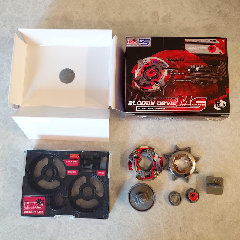 May include: A boxed toy set with various components, including a red, black, and silver spinning top, a metal ring, and a black plastic case. The box features the text "Bloody Devil MS" and "Standard Version".