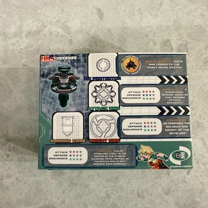 May include: A boxed toy set featuring a green and black spinning top with a detailed design. The packaging includes text and graphics highlighting the toy's features, including attack, defense, and endurance ratings. The box also has the text "HM2 DEFENSE".