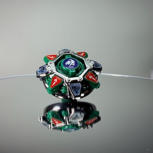 May include: A green, red, silver, and black spinning top toy. The top has a circular design with a central green core and a silver outer ring. Red and blue accents are visible. The toy is reflected on a mirrored surface.