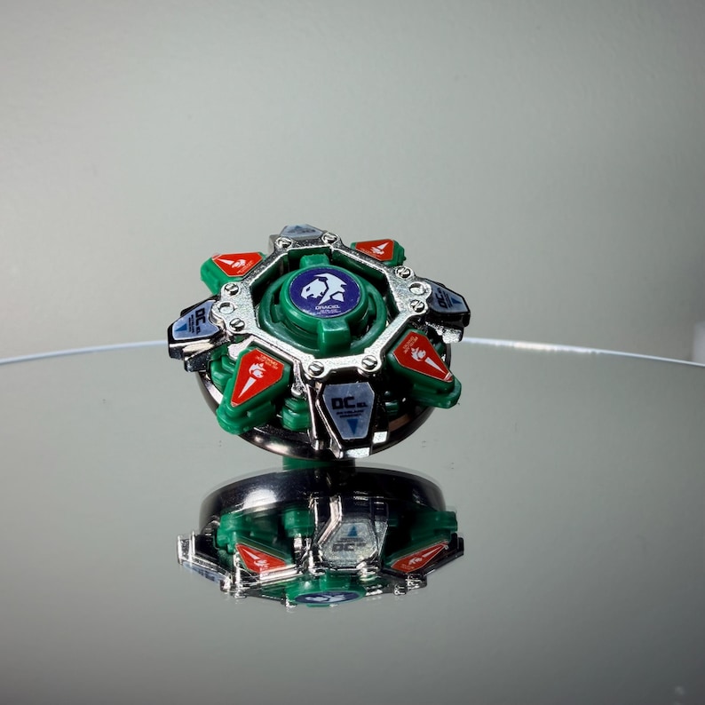May include: A green, red, silver, and black spinning top toy. The top has a circular design with a central green core and a silver outer ring. Red and blue accents are visible. The toy is reflected on a mirrored surface.