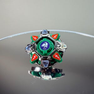 May include: A green and silver toy with red accents. The toy has a circular design with a central emblem and text. The toy is sitting on a reflective surface.