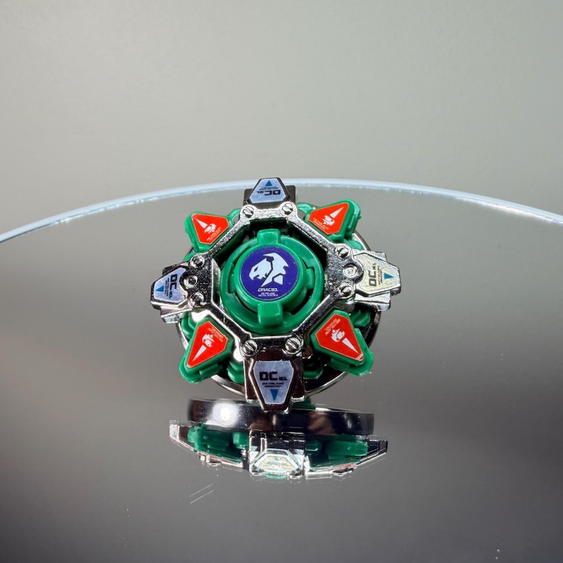 May include: A green and silver toy with red accents. The toy has a circular design with a central emblem and text. The toy is sitting on a reflective surface.