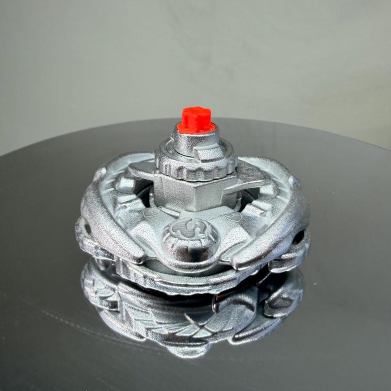 Puede incluir: Un trompo de color plateado con un bot&oacute;n central rojo. El juguete tiene un dise&ntilde;o complejo y multicapa con detalles intrincados. La parte superior est&aacute; dise&ntilde;ada para girar sobre una superficie plana.