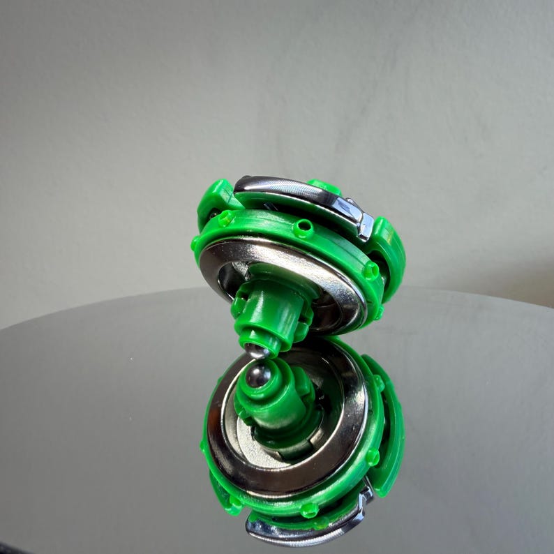 Pu&ograve; includere: Un giocattolo trottola verde e argento. Il giocattolo ha un design circolare con un esterno in plastica verde e un interno in metallo argentato. Il giocattolo &egrave; appoggiato su una superficie riflettente.