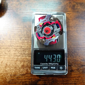 May include: A colorful spinning top toy with red, gray, and blue accents rests on a digital scale. The scale displays a weight of 44.30 grams. The toy has a circular design with intricate details and a central emblem.