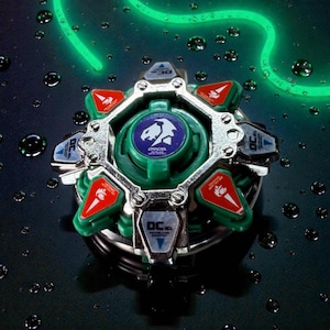 May include: A green and silver toy with red and blue accents. The toy has a central green disc with a white and blue emblem and the word "DRACIEL" printed on it. The toy is surrounded by silver metal and has several red and blue triangular pieces.