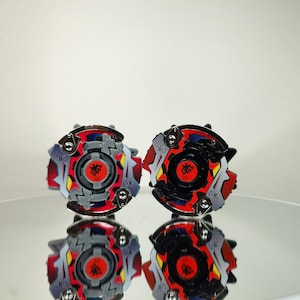 May include: Two colorful spinning tops with red, gray, black, and yellow accents. Each top features a central red emblem and a complex, multi-layered design. The tops are displayed on a reflective surface.