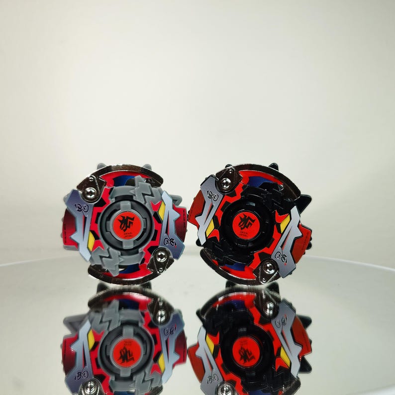 May include: Two colorful spinning tops with red, gray, black, and yellow accents. Each top features a central red emblem and a complex, multi-layered design. The tops are displayed on a reflective surface.
