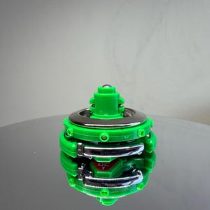 Pu&ograve; includere: Una trottola verde e argento. Il giocattolo ha un corpo verde brillante con dettagli argentati e una piccola sfera argentata sulla parte superiore. Il giocattolo ha un logo DC.