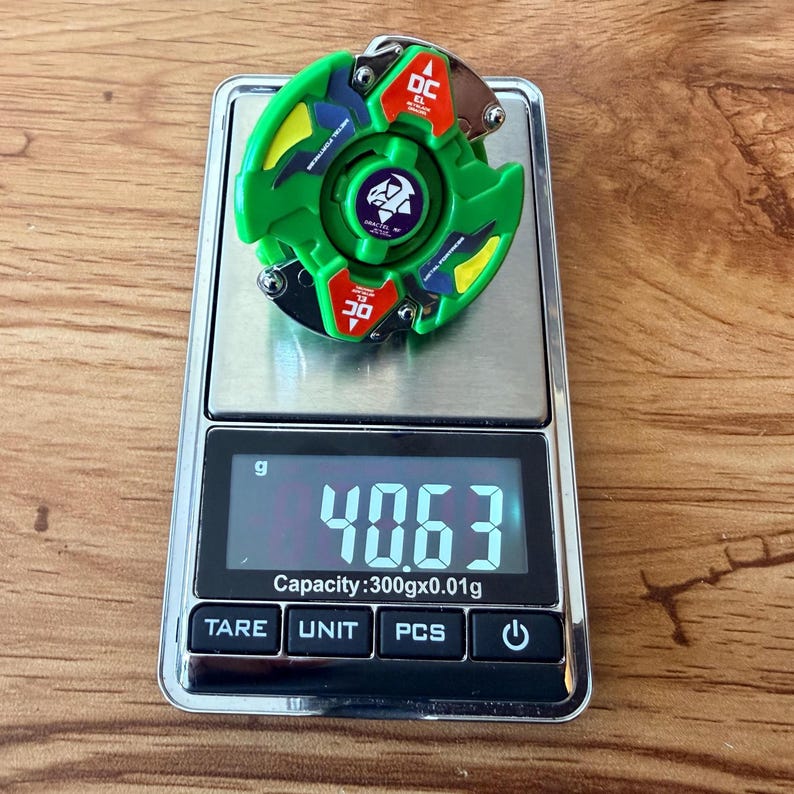 Pu&ograve; includere: Un giroscopio giocattolo verde e rosso con un emblema viola &egrave; posto su una bilancia digitale. La bilancia mostra un peso di 40,63 grammi. Il giocattolo ha dettagli gialli e blu.